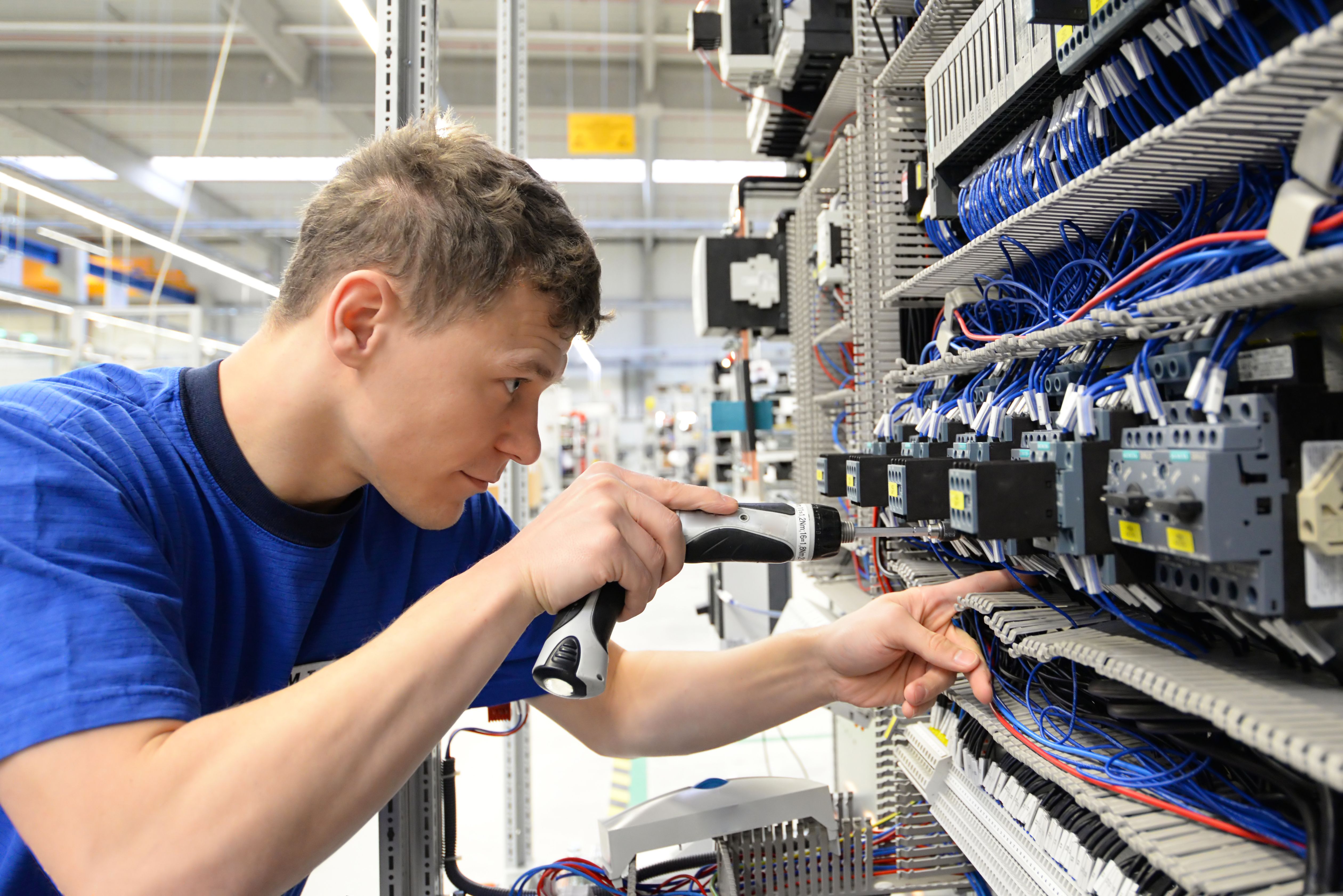 Monteur arbeitet an einem Schaltschrank und verbindet Kabel in einer industriellen Produktionshalle.
