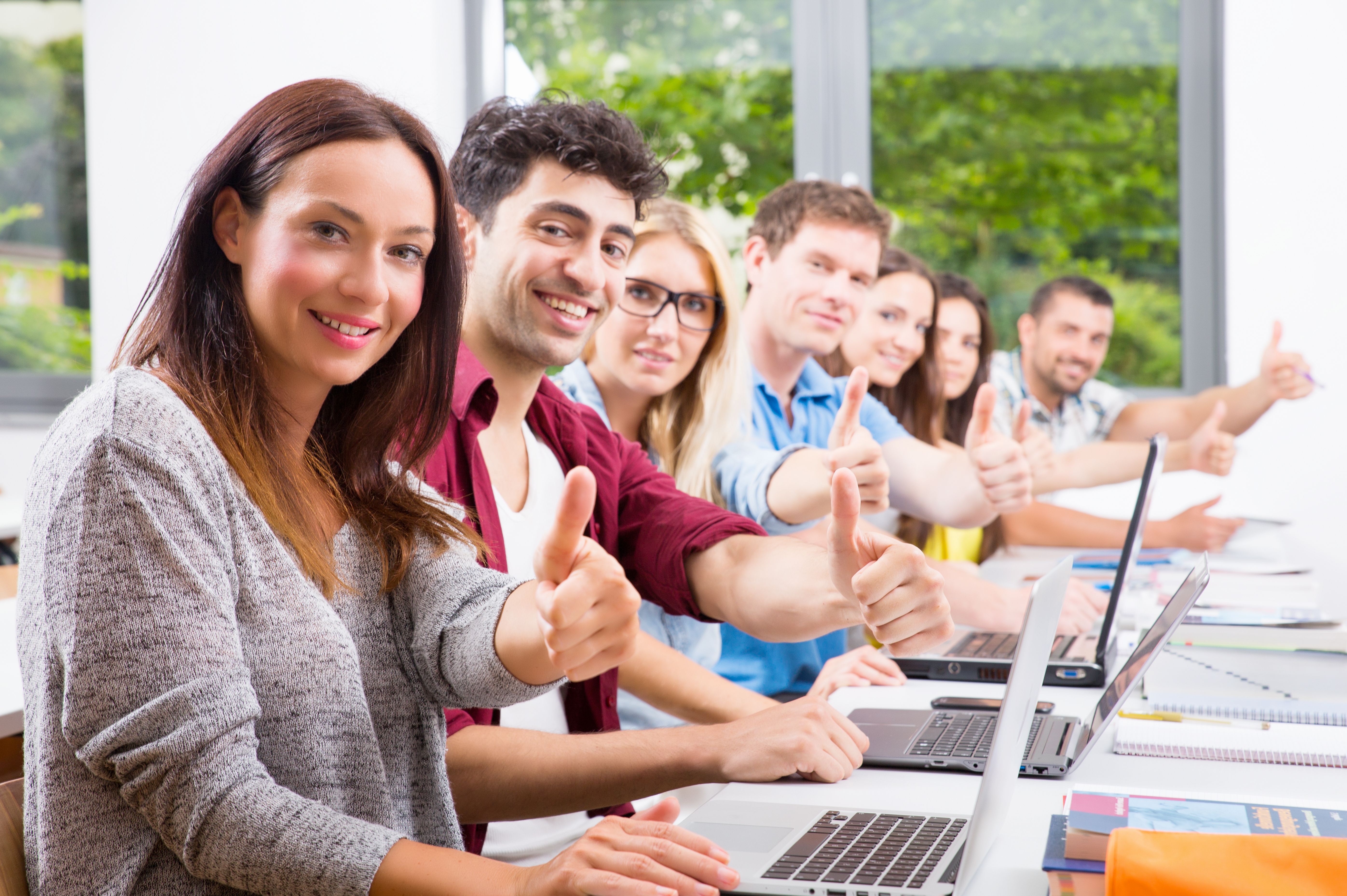 Teilnehmende einer Weiterbildung sitzen im Seminarraum mit Laptops und zeigen Daumen hoch für erfolgreichen Kursabschluss